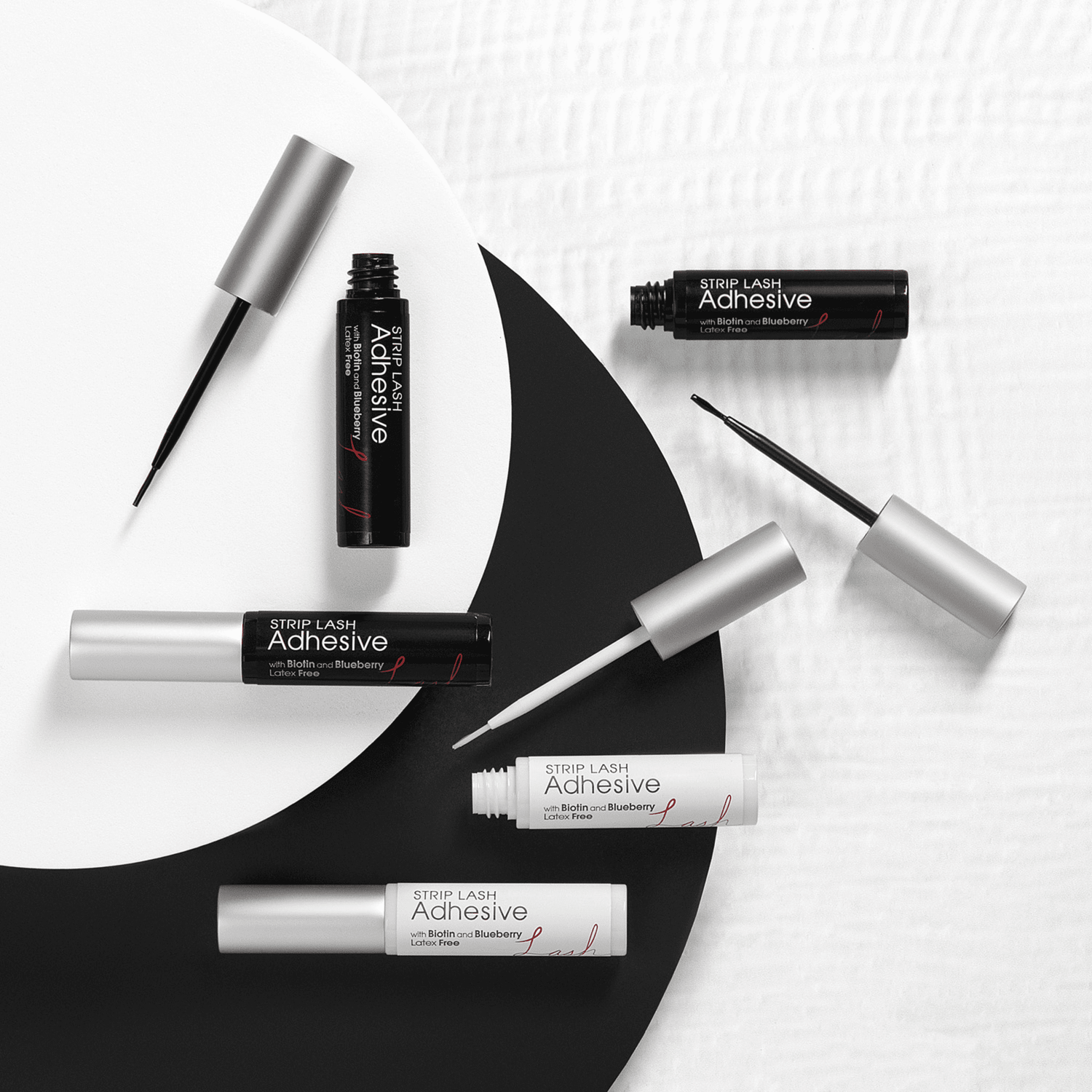 Six tubes of KISS Strip Lash Adhesive in black and white packaging with silver caps, some open to show fine applicator brushes; labeled latex-free with biotin and blueberry.