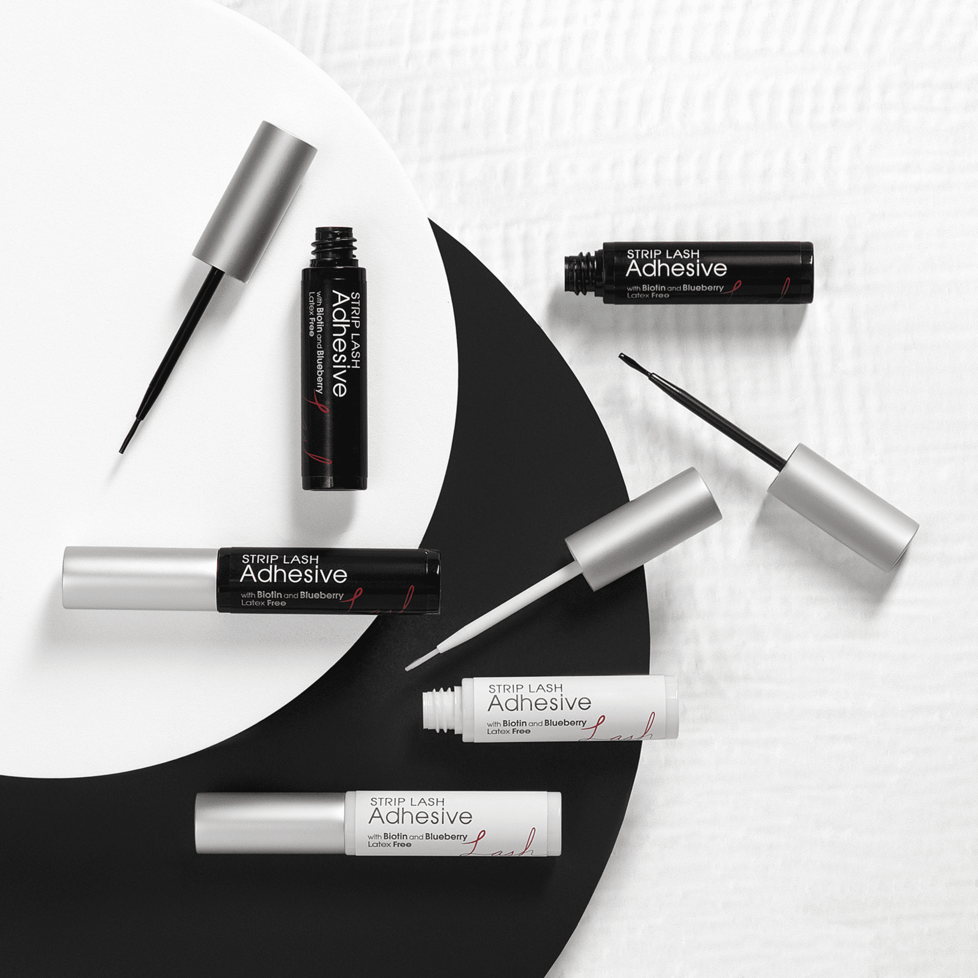 Six tubes of KISS Strip Lash Adhesive in black and white packaging with silver caps, some open to show fine applicator brushes; labeled latex-free with biotin and blueberry.