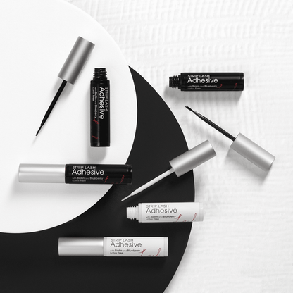 Six tubes of KISS Strip Lash Adhesive in black and white packaging with silver caps, some open to show fine applicator brushes; labeled latex-free with biotin and blueberry.