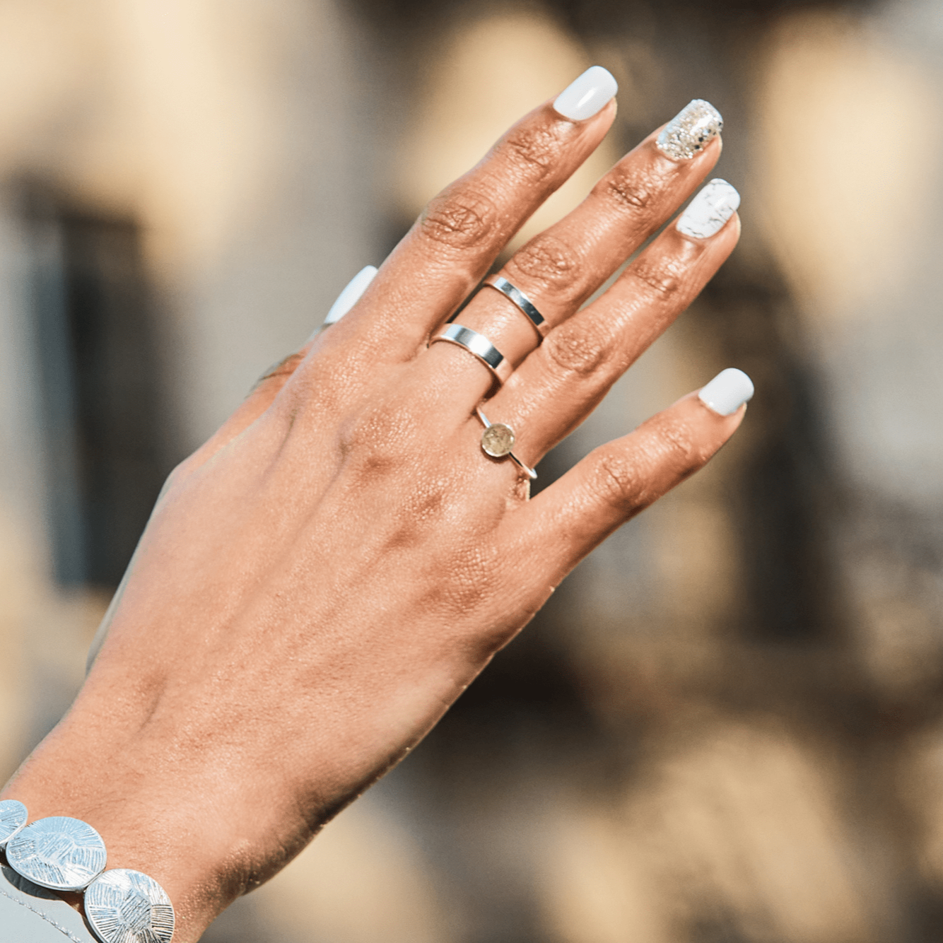 Hand with light brown skin wearing silver rings and a silver bracelet, featuring white press-on nails with one glittery silver nail and one white nail with delicate gray marbling.