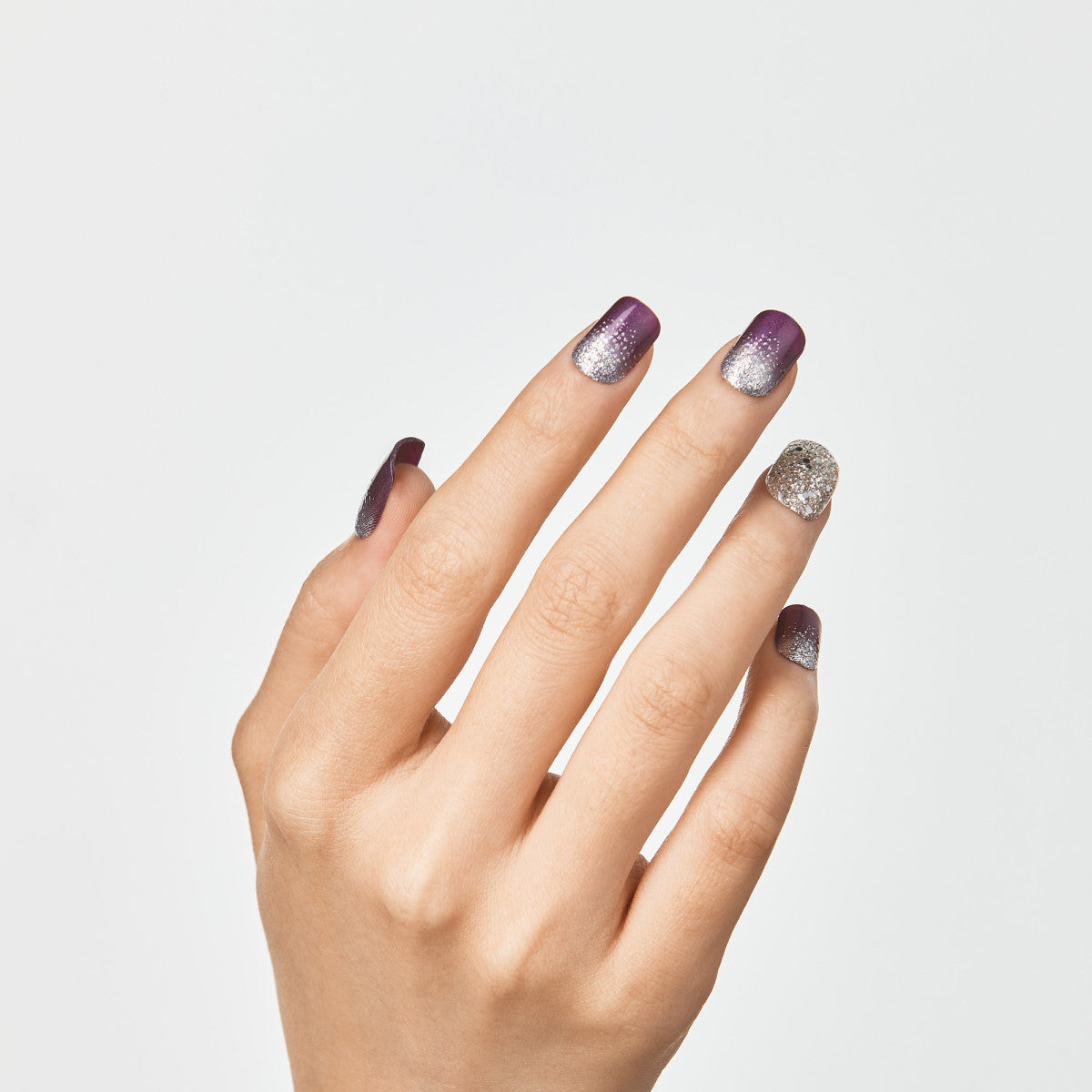 Hand with five nails wearing imPRESS press-on manicure in Heartquake design, featuring purple to silver glitter ombré on four nails and full silver glitter on the ring finger.