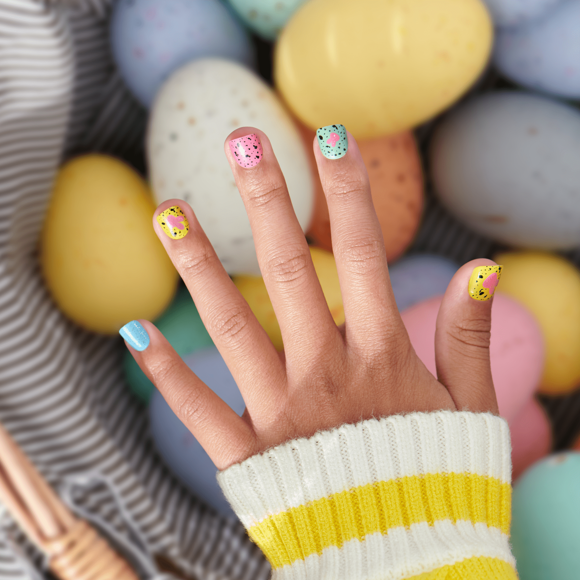 Pastel Easter egg nail art with colorful speckles on hand against Easter eggs background.