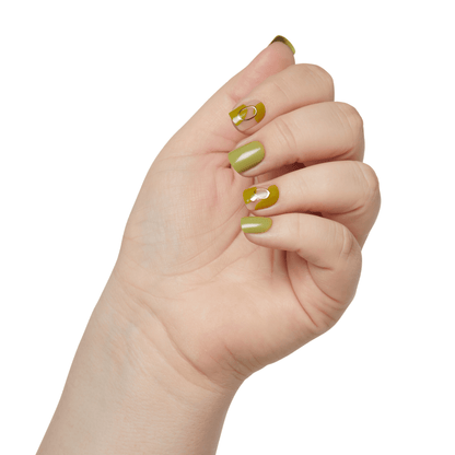 Hand with press-on nails in olive green shades; some nails have a glossy half-moon design with a gold metallic ring accent.