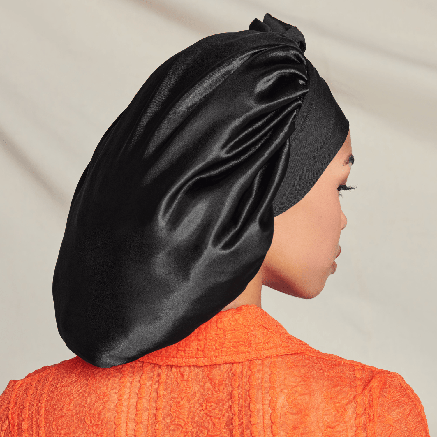 Person in profile wearing black silk bonnet and orange textured shirt against neutral background.
