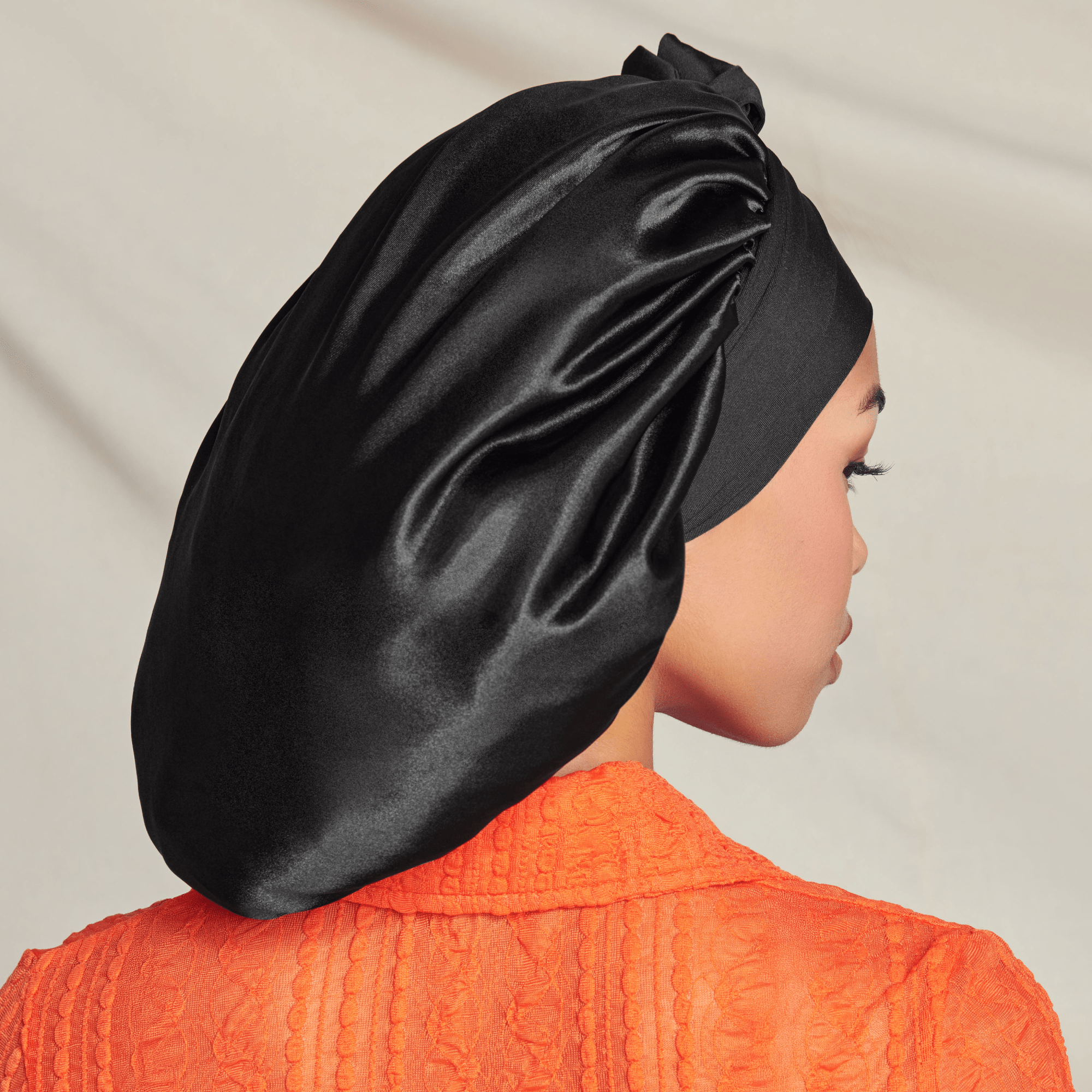 Person in profile wearing black silk bonnet and orange textured shirt against neutral background.