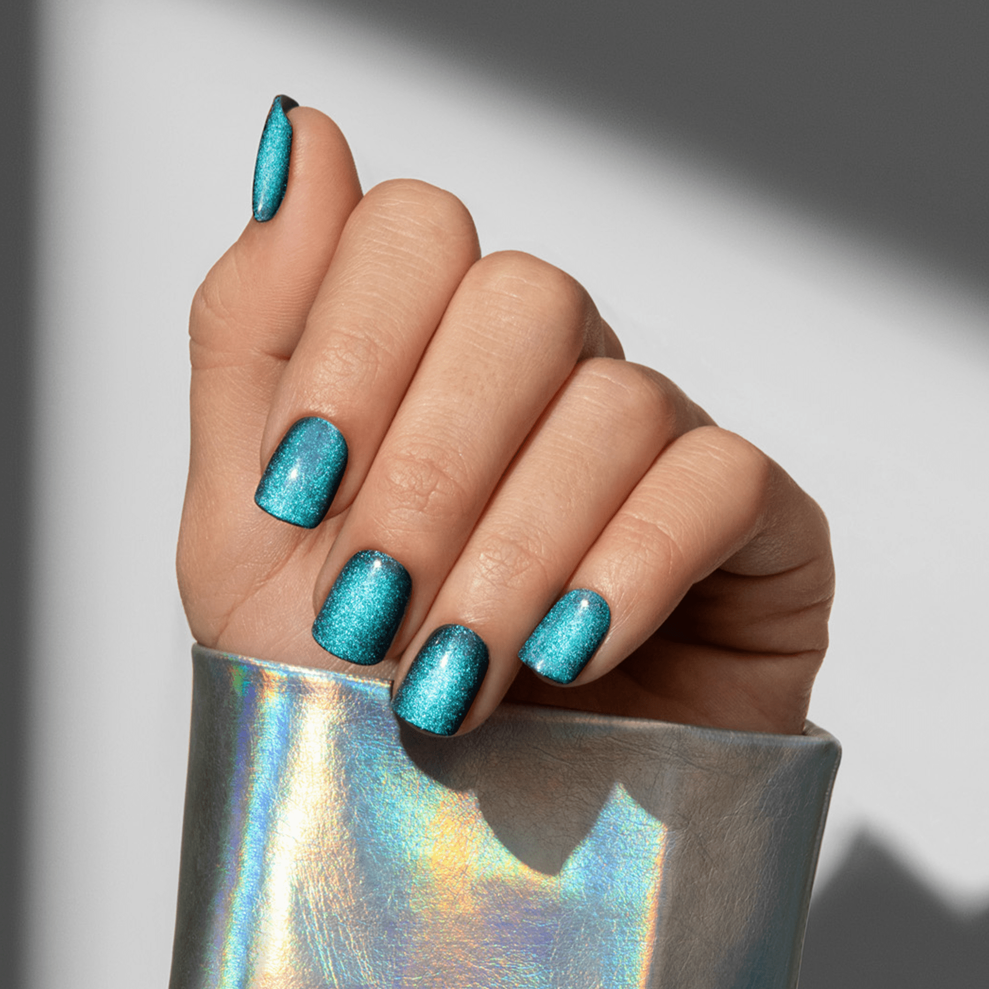 Hand with press-on nails in shimmering turquoise blue color, nails are medium length with rounded square shape, shown against a light background, wearing a metallic holographic silver sleeve.